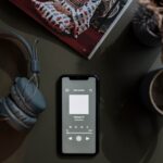 A flat lay pic of a mobile play one of the best event podcasts, kept aside a pair of headphones and a cup of coffee, all of which are kept atop a table
